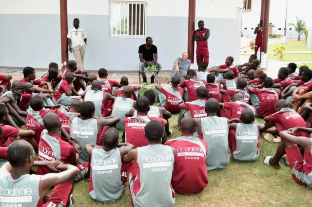 Young players aged 12 to 14 sitting on the ground listening to coaches during a training session.